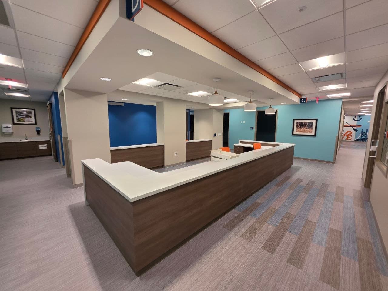 Completed nurse station: L-shaped casework with a solid-surface top, soffit with an orange reveal, pendant lights over the corridor beyond