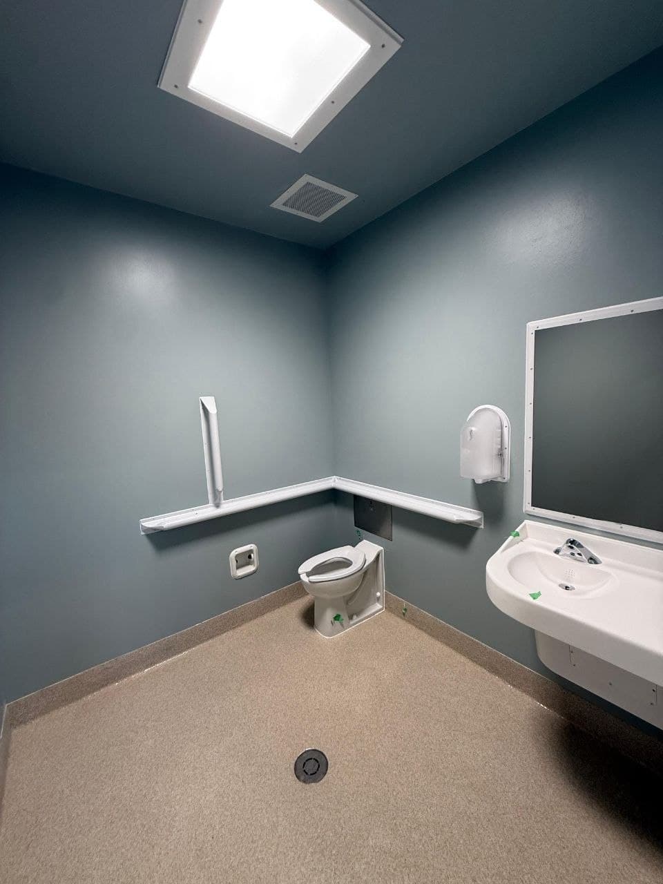 Completed restroom with ligature-resistant fittings: sloped grab bar and shelf, security mirror, seamless resinous floor coved to the wall